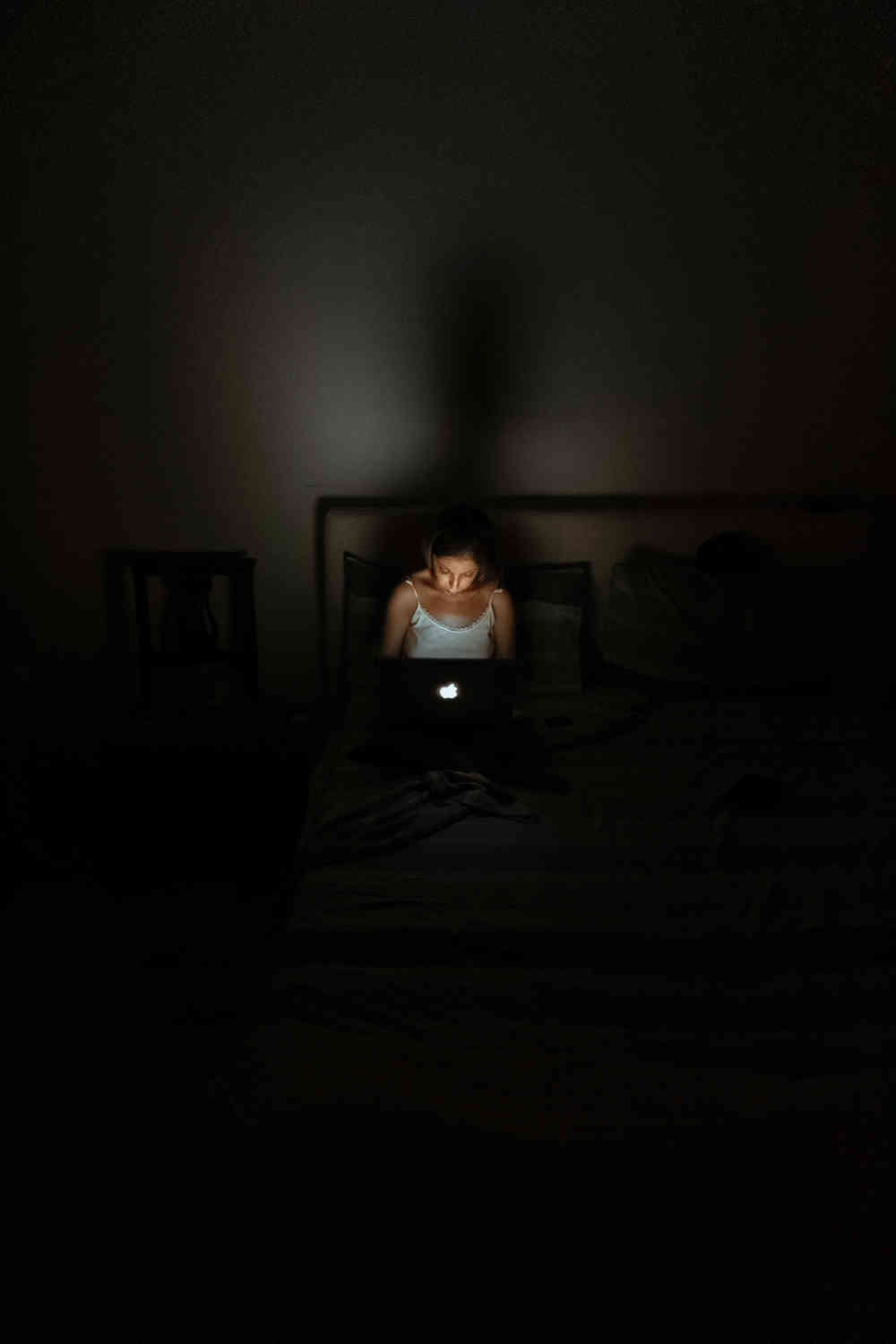 Woman sitting in the dark looking at an open Macbook that has its illuminated Apple logo on the lid.